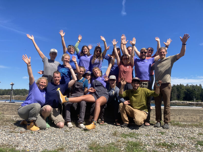 The image shows a group of people posing outdoors on a sunny day. They are raising their hands in the air, smiling, and appear to be enjoying themselves. The background includes a clear blue sky and some trees. It looks like a team or group of friends celebrating something.